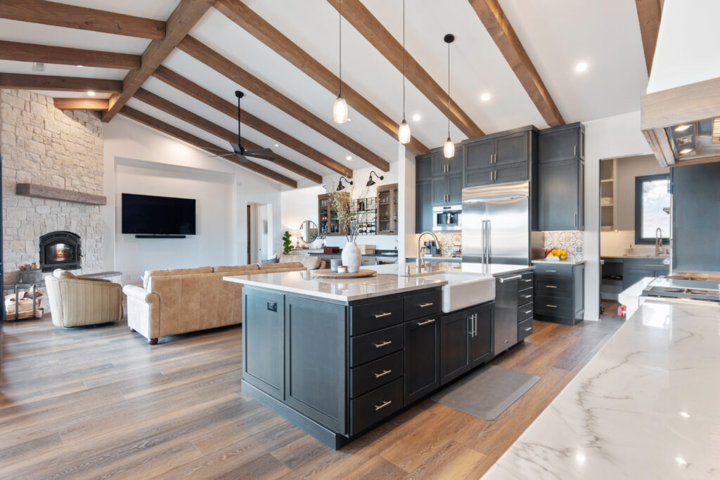 Open-floor-plan-steel-frame-barndominium-designs-home-interior-with-faux-beams-ceiling-wall-feature-stone-fireplace-and-wide-plank-wood-floors
