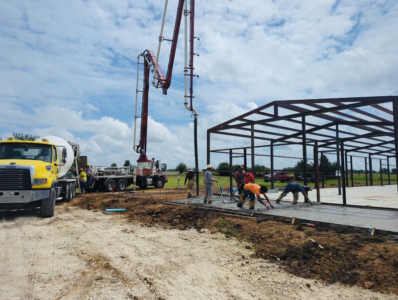 Metal building site prep showing a pump truck on site with a crew leveling concrete and a custom metal building erected in the background.